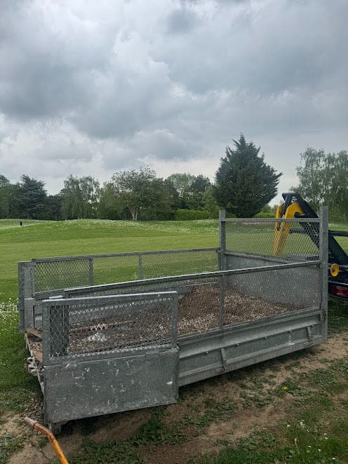 Benne à ridelles grillagées chargée de terre et de végétaux, posée en bordure d'un grand espace vert arboré